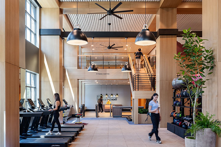 The interior of the fitness center shows a row of treadmills near several large windows. One person is walking on one of the treadmills, and another person is walking across the room.