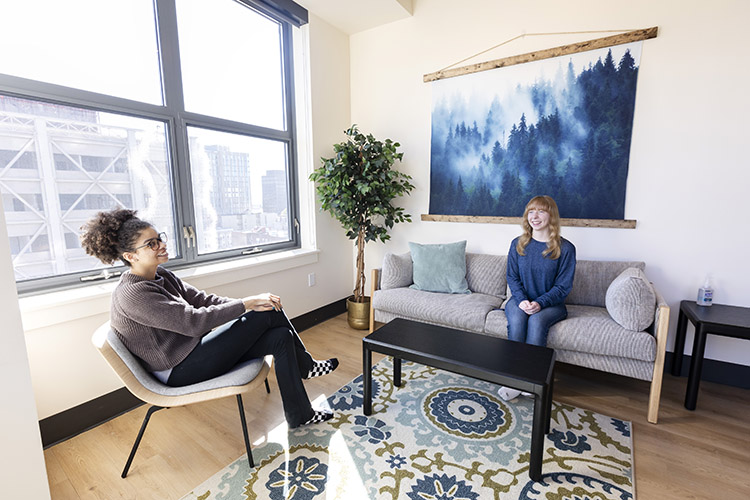 Two students sit in a furnished living room. One student is sitting on a couch, looking at the other student, who is sitting in an armchair across the room.