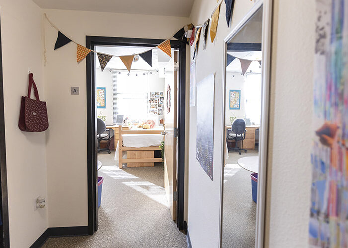 An empty hall way with a wall-hanging mirror on the right side leads to two doors to other rooms. One of the rooms is a bedroom, and the bed and desk are partially visible.