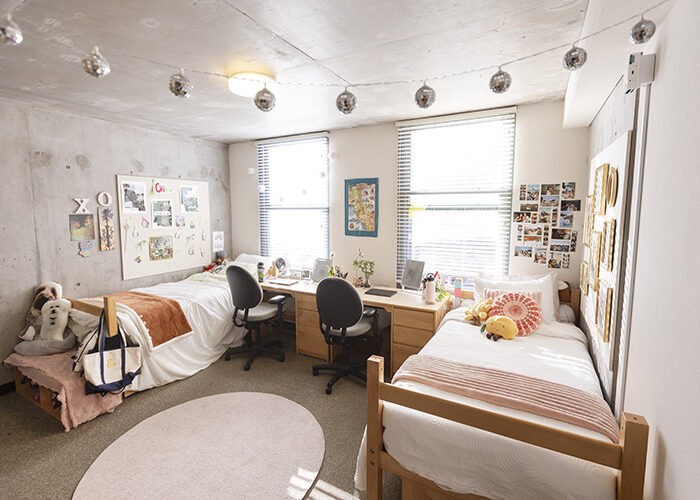 A well-lit double occupancy bedroom features two twin beds across from each other on opposite sides of the room. There are two desk chairs and two wooden desks underneath two large windows adjacent to the beds.