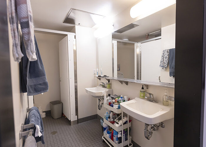 An open door displays an empty shared bathroom, with two sinks underneath a large mirror and towels hanging on the opposite wall.