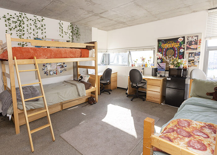 An empty triple-occupancy bedroom is fully-furnished and decorated. One twin bed sits against the wall on the right side of the room, and a bunk bed is positioned on the opposite side of the room. In the back of the room, three desks and a mini-fridge sit next to each other, positioned under several windows.