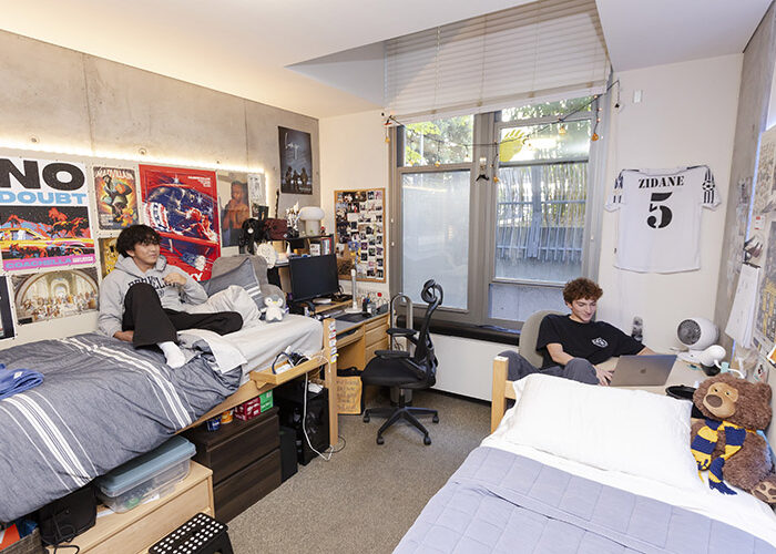 Two students sit in a double-occupancy mini-suite bedroom. One student sits on a bed on the left side of the room, while the other student sits at a desk with a laptop on the other side of the room. There are many posters and decorations on the walls above each bed.