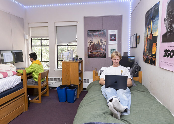 An interior shot of a double room in Foothill shows one student working at a desk, and one student working on a laptop while lying in a twin-sized bed.