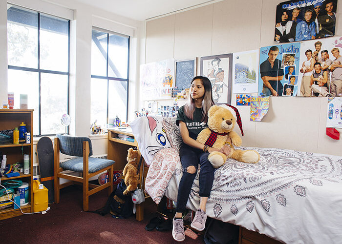 An interior shot of a bedroom shows a student sitting on a twin-sized bed next to large windows. The room is decorated with posters and photos on the wall.