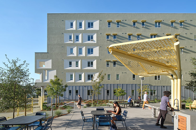 A graduate student with a laptop sits at an outdoor table in a courtyard. Another student washes his hands at a nearby concrete sink. There are other tables behind them, as well as an outdoor barbecue area.