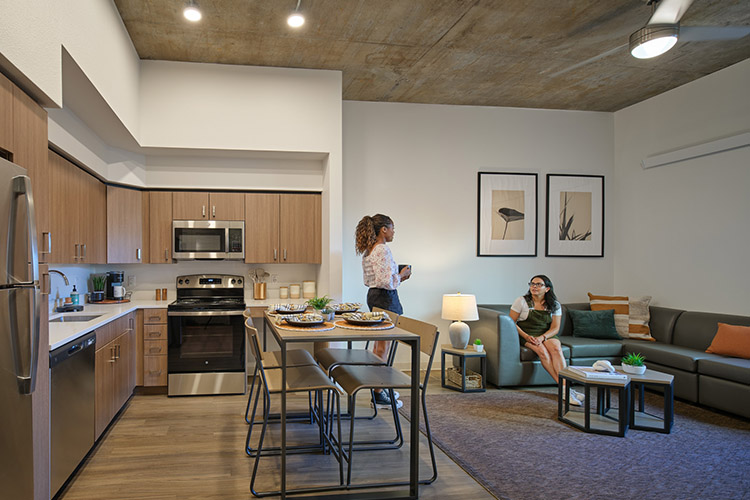 The kitchen of a 2-bedroom apartment is shown with many appliances and a kitchen table. One student stands near the kitchen, talking to another student who is sitting on a sectional couch in the living room area.