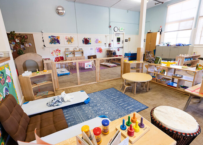 The interior of an indoor child care center is shown featuring musical instruments, chairs and tables, and children’s art projects.