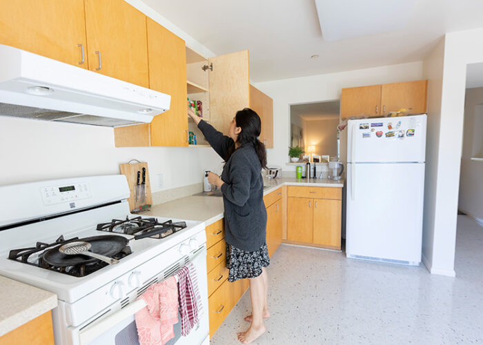 A woman stand in the kitchen next to the stove, reaching into a cabinet.